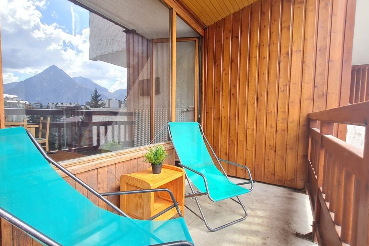 Two bright turquoise deck chairs on a wooden balcony with a small side table and a view of mountains.
