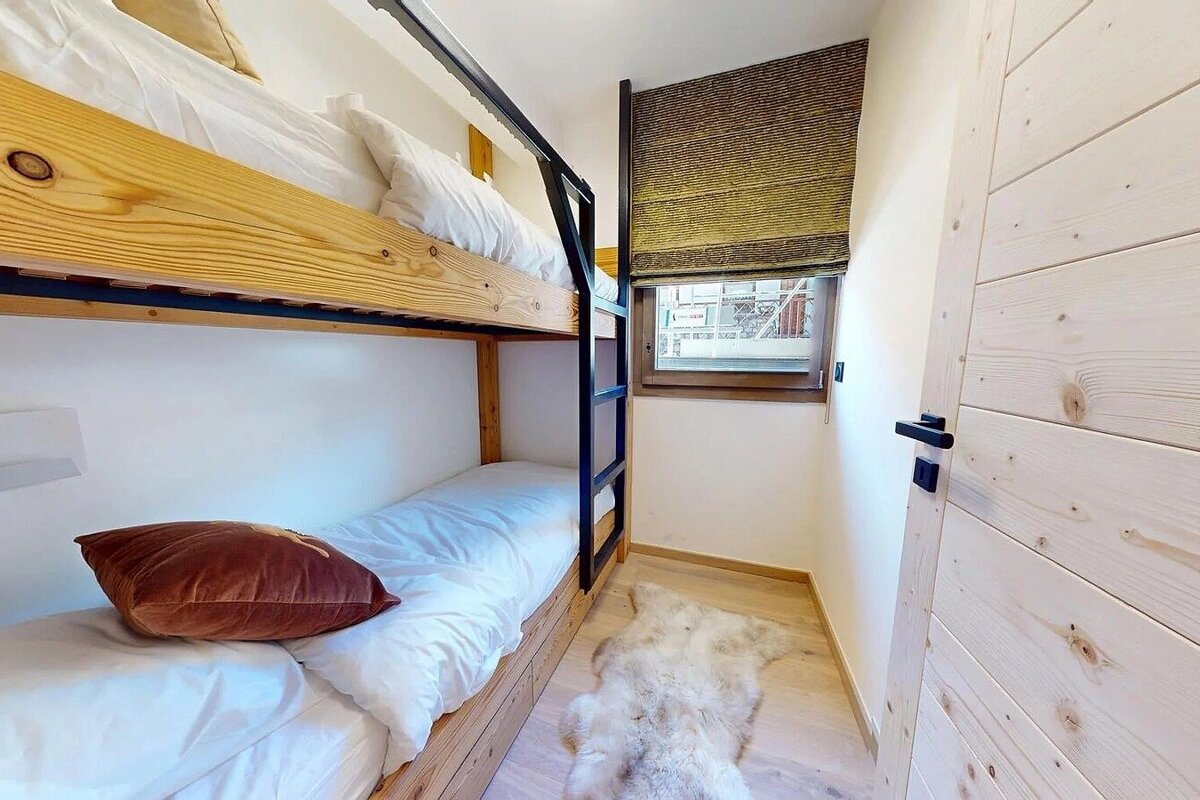 Cozy room with wooden bunk beds, white bedding, a window with a textured blind, and a fluffy white rug on light wood flooring.