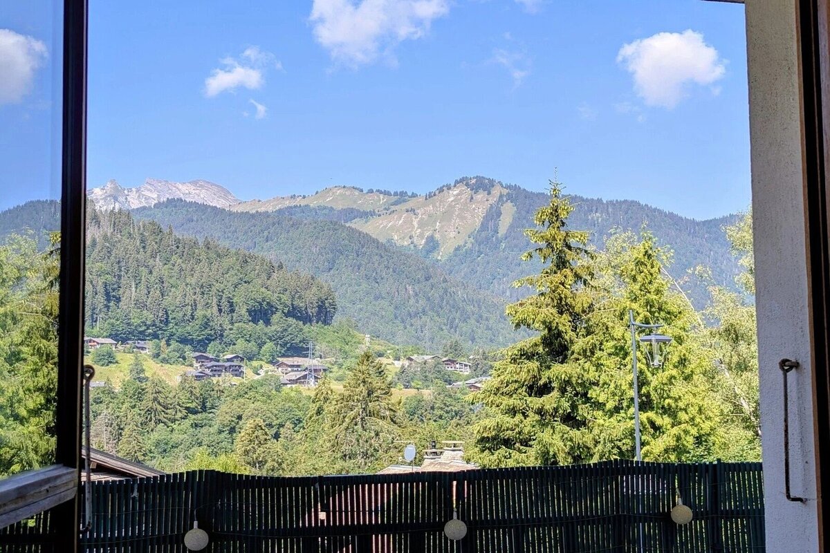 Scenic view of forested mountains and a small alpine village under a blue sky, framed by a window and balcony fence.