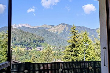 Scenic view of forested mountains and a small alpine village under a blue sky, framed by a window and balcony fence.