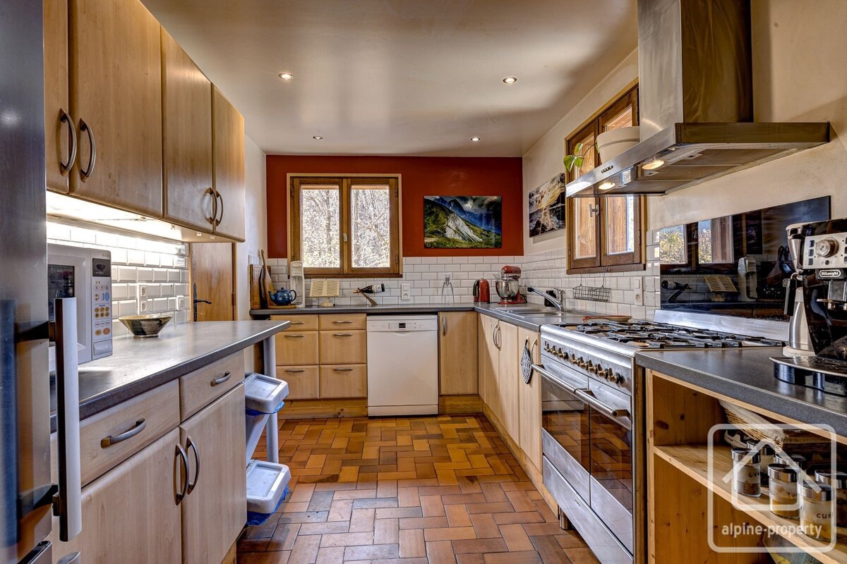 A modern kitchen with light wood cabinets, a red accent wall, white subway tile backsplash, stainless steel appliances, and a tiled floor.