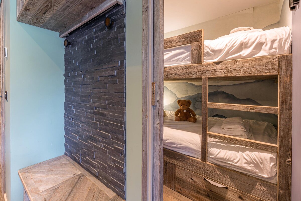 A rustic bedroom with wooden bunk beds and a teddy bear, viewed from a hallway with a dark stone accent wall.