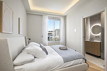Bright, modern bedroom with a large bed, city-view window, and an en-suite bathroom featuring an illuminated round mirror.