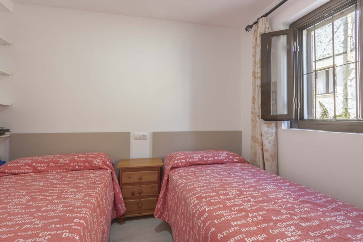 A twin bedroom features two beds with red, text-patterned bedspreads, a wooden nightstand between them, and an open window.