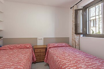 A twin bedroom features two beds with red, text-patterned bedspreads, a wooden nightstand between them, and an open window.