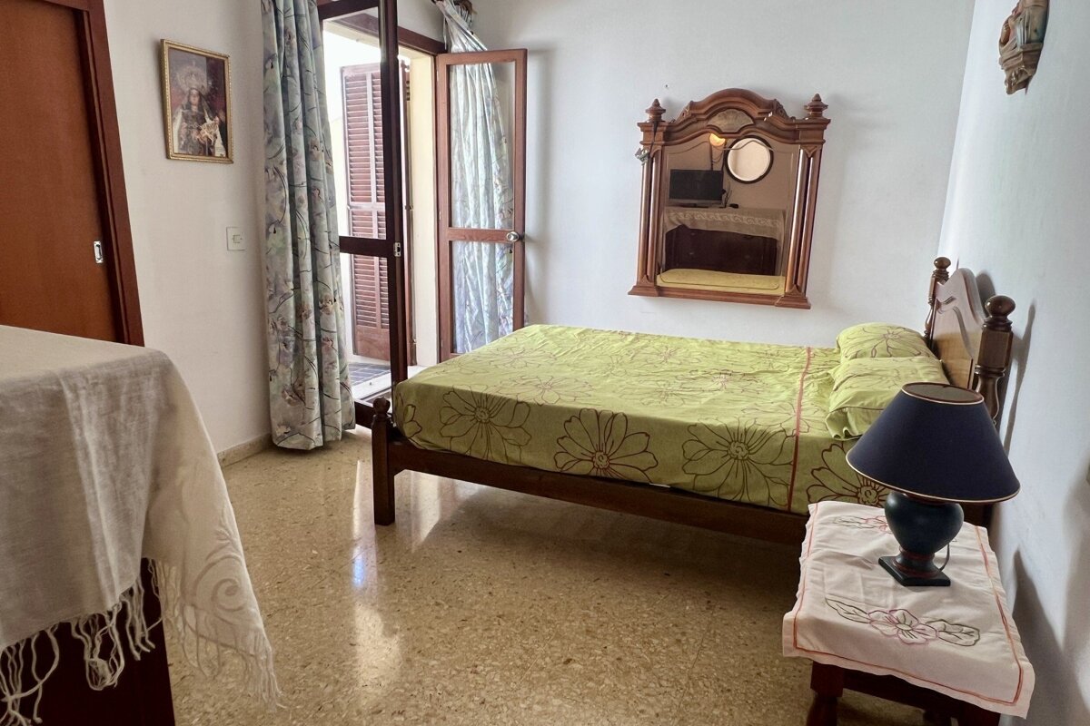A bedroom with a wooden bed, green floral bedding, and a decorative mirror above it. An open door with shutters leads outside, and a nightstand with a lamp sits beside the bed.