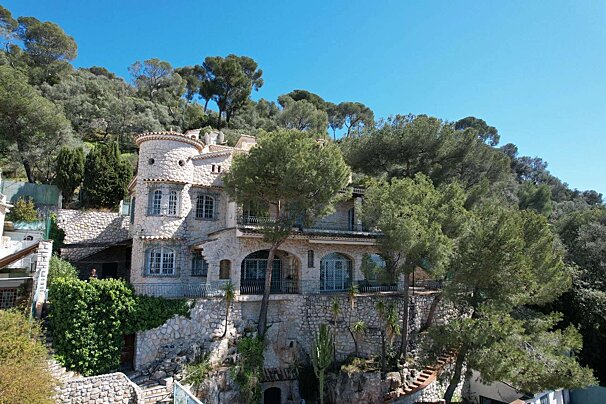 A multi-story stone house with a round tower built into a lush green hillside under a clear blue sky, surrounded by tall trees.