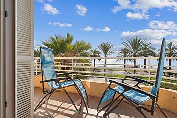 Balcony with two blue chairs, offering a view of palm trees, the ocean, and a clear blue sky with fluffy white clouds.
