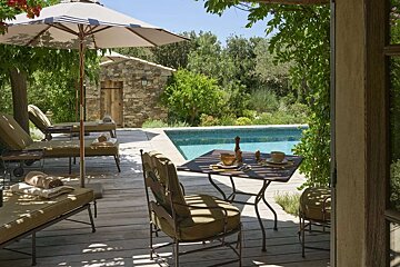 An inviting outdoor space featuring a swimming pool, wooden deck with lounge chairs and dining set, lush landscaping, and a rustic stone building.