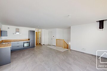 An empty, modern open-plan room with grey tiled flooring, a fitted kitchen, and a wooden staircase leading downstairs.