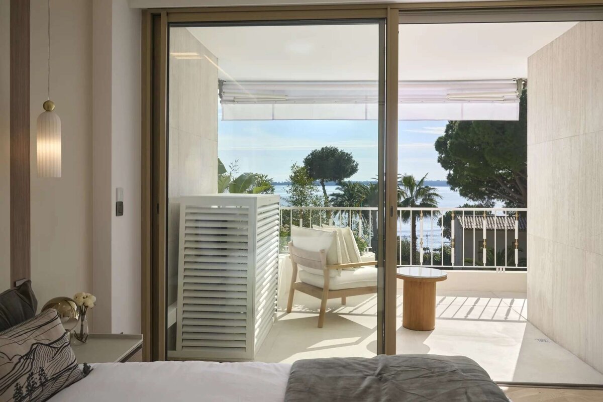 A bright bedroom with sliding doors opening to a balcony featuring an armchair, louvered unit, and a beautiful sea view framed by trees.
