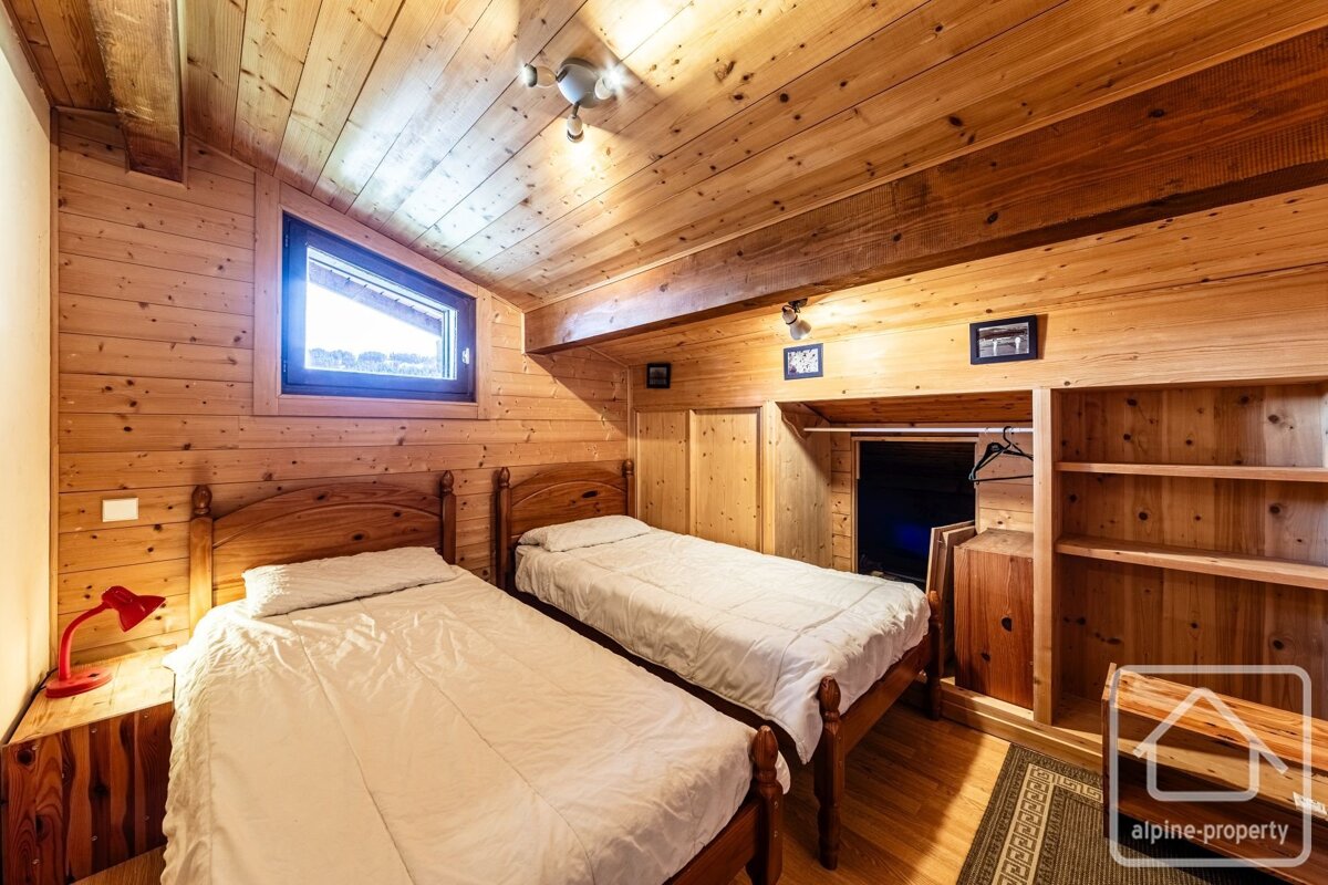 A cozy, rustic bedroom fully paneled in wood, featuring two single beds, a slanted window, and built-in shelves/wardrobe.