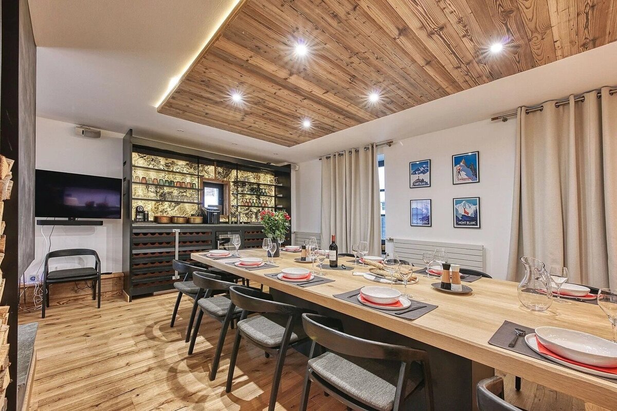 A modern dining room with a long wooden table set for dinner. Features a wooden plank ceiling, ski posters, a bar area, and a wall-mounted TV.