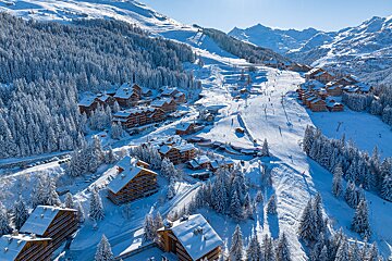Aerial view of a snowy ski resort in the mountains, with chalets, snow-covered pine trees, ski slopes, and skiers under a clear blue sky.