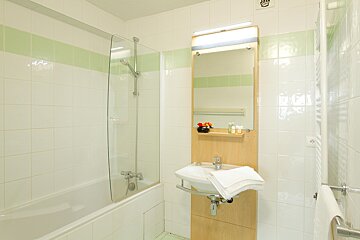 A clean, white-tiled bathroom with a green stripe, featuring a bathtub with a glass shower screen, a sink with a mirror and wooden vanity, and fresh towels.