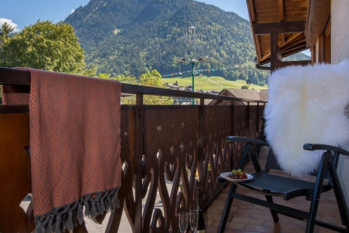 Cozy wooden balcony with a patterned blanket and a white fur throw-draped chair, overlooking beautiful green mountains.