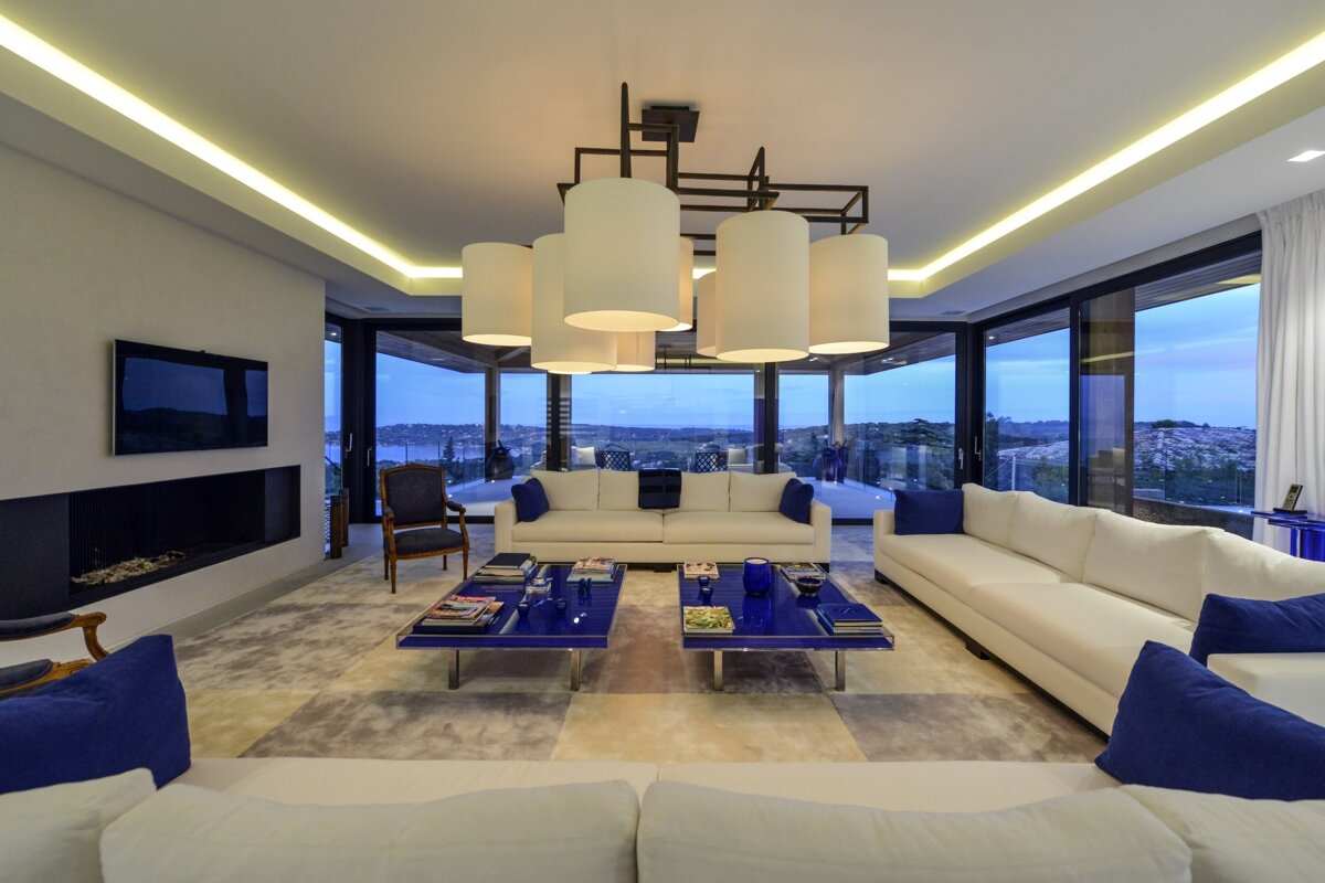 Luxurious, modern living room with white sofas, vibrant blue accents, large scenic windows, sleek fireplace, and a stylish chandelier.