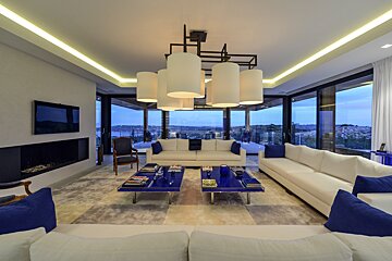Luxurious, modern living room with white sofas, vibrant blue accents, large scenic windows, sleek fireplace, and a stylish chandelier.