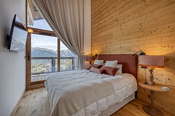 A warm, wood-paneled bedroom with a large window and balcony overlooking a scenic mountain town. Features a plush bed, TV, and cozy lighting.