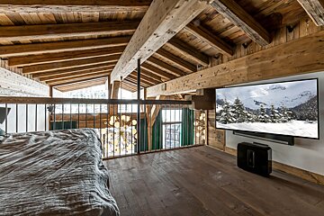 Rustic wooden chalet loft with a bed, railing, and a large TV showing a snowy mountain landscape, complemented by exposed beams and floor.