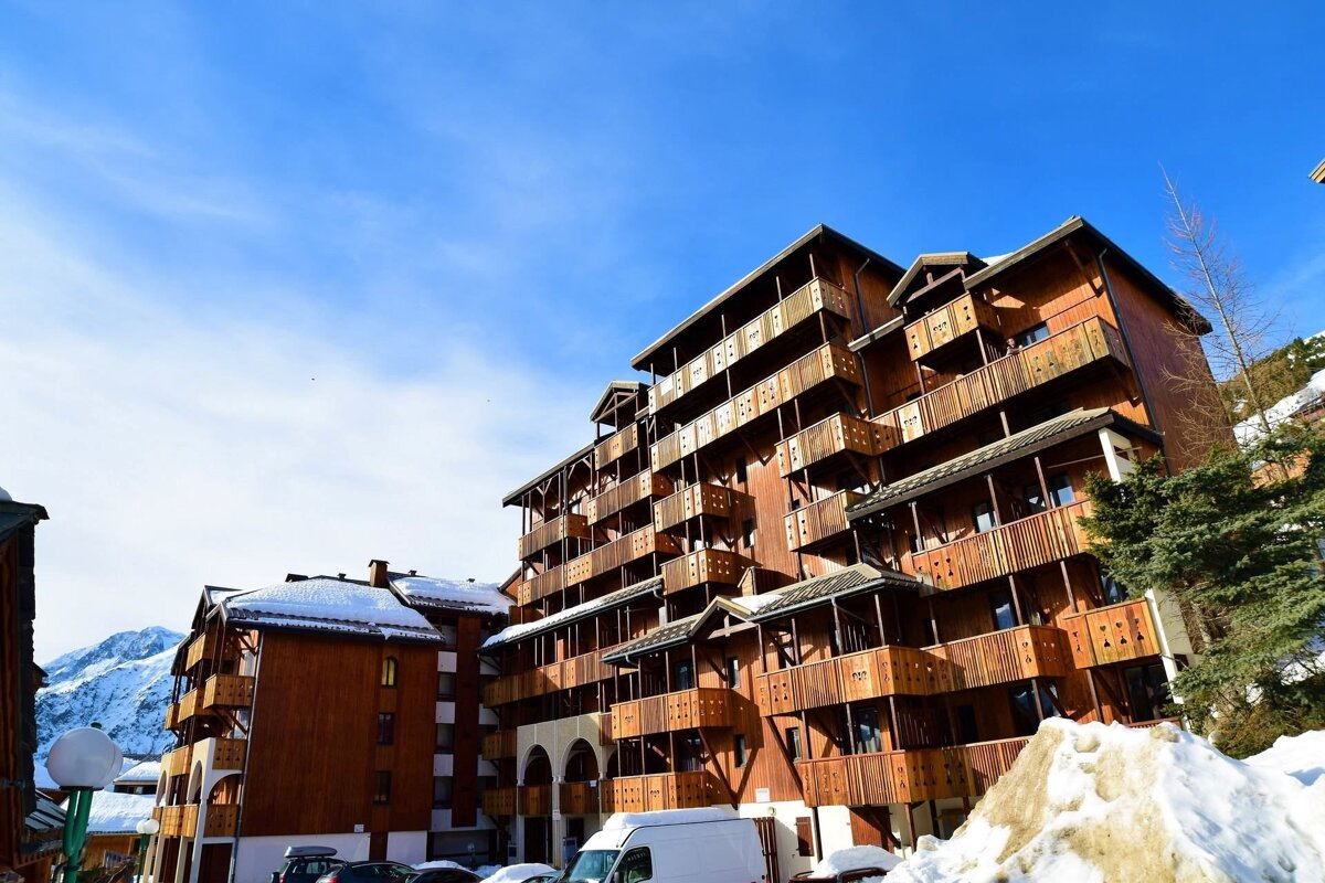 A multi-story wooden chalet-style hotel or apartment building with many balconies, set against a bright blue sky with snow on the ground in a mountain resort.