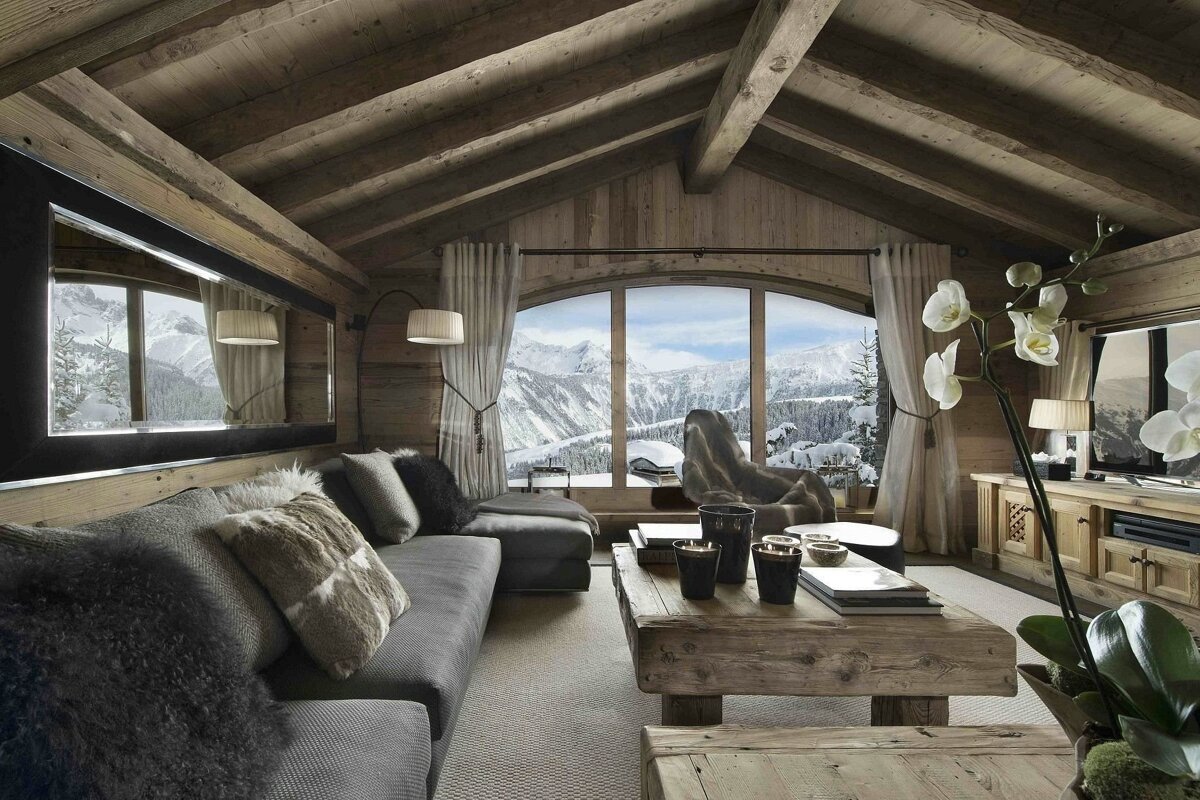 Cozy wooden chalet living room with an arched window showcasing snowy mountains. A gray sectional, rustic coffee table, and orchid plant create a luxurious ski lodge feel.