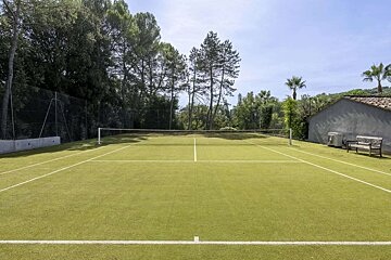 A vibrant green outdoor tennis court with white lines, surrounded by tall trees. A small building and a wooden bench are visible on the right.