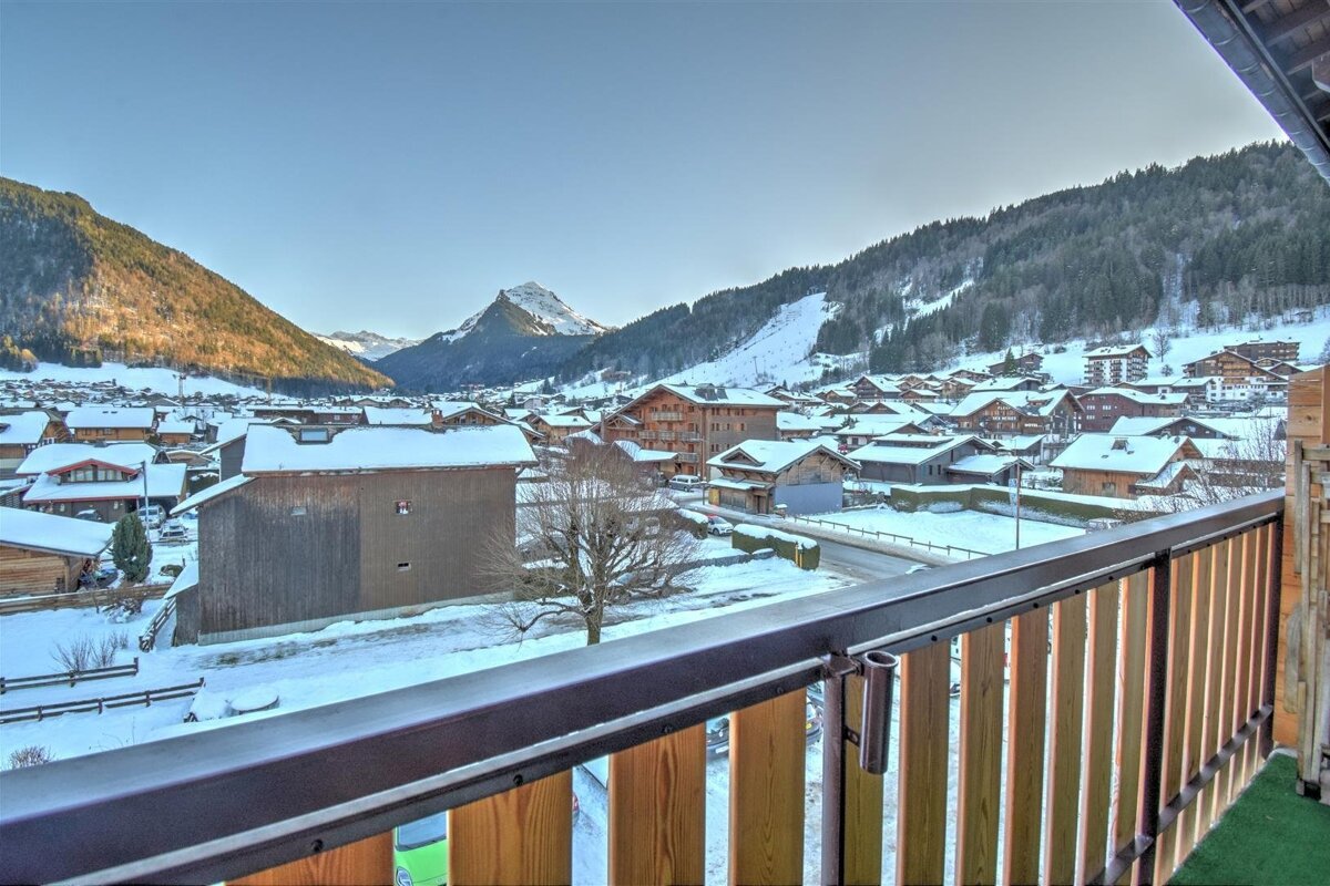 A scenic balcony view of a snow-covered alpine village nestled in a mountain valley with wooden chalets, surrounded by forested slopes under a clear sky.