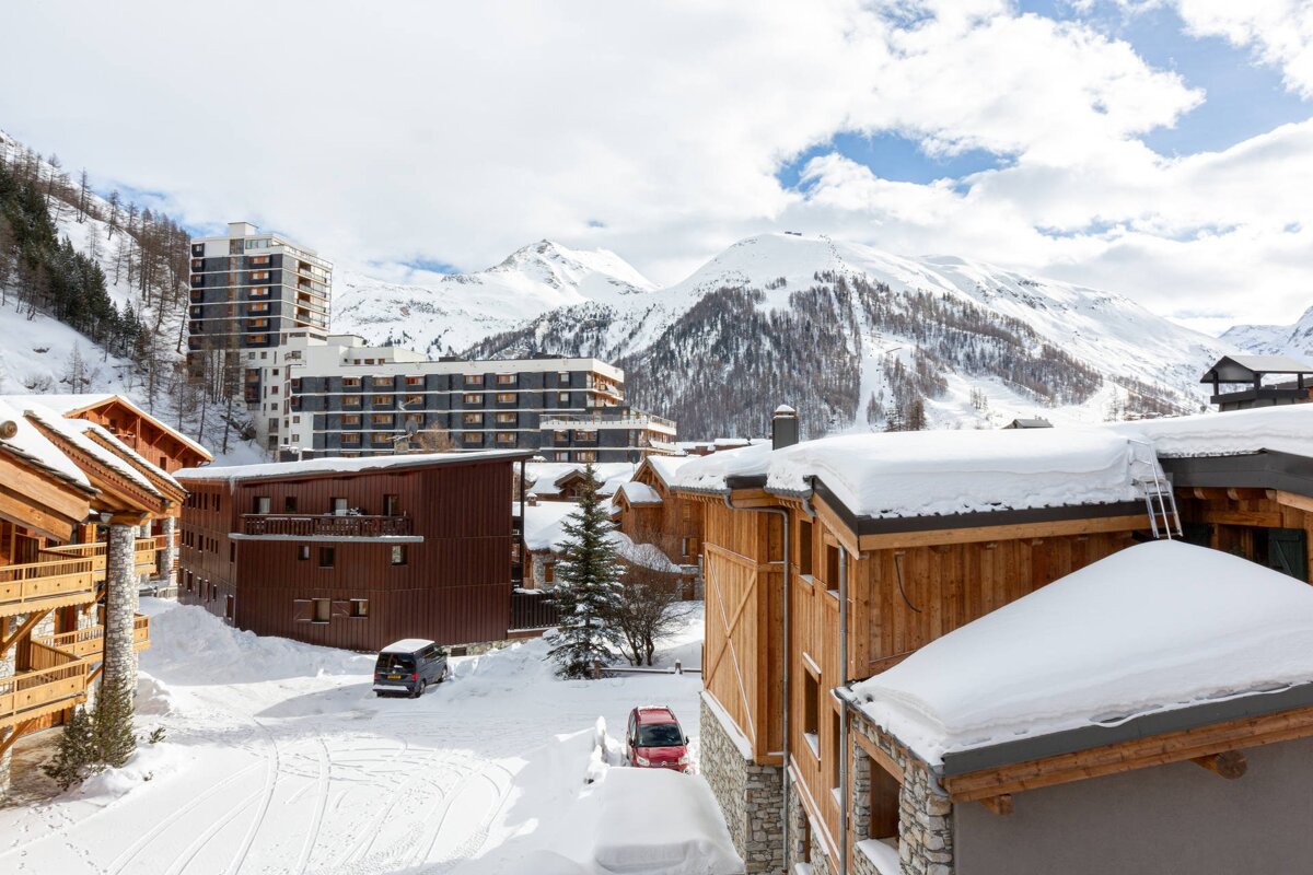 Scenic winter view of a snowy mountain village with wooden chalets, modern buildings, and snow-capped peaks in the background under a cloudy sky.