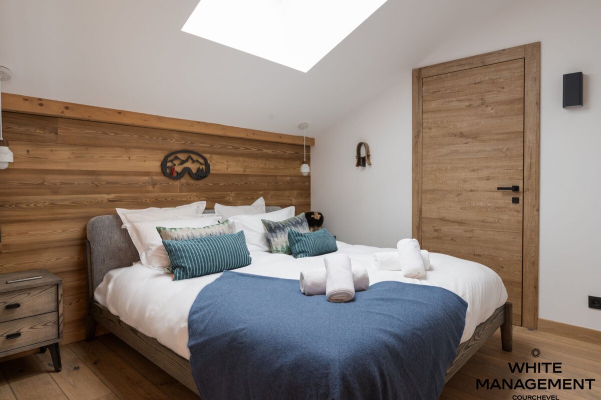 A cozy, modern bedroom featuring a large bed with white linens and a blue throw. It has a wooden headboard, floor, and door, a skylight, and ski-themed decor.
