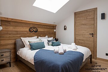 A cozy, modern bedroom featuring a large bed with white linens and a blue throw. It has a wooden headboard, floor, and door, a skylight, and ski-themed decor.