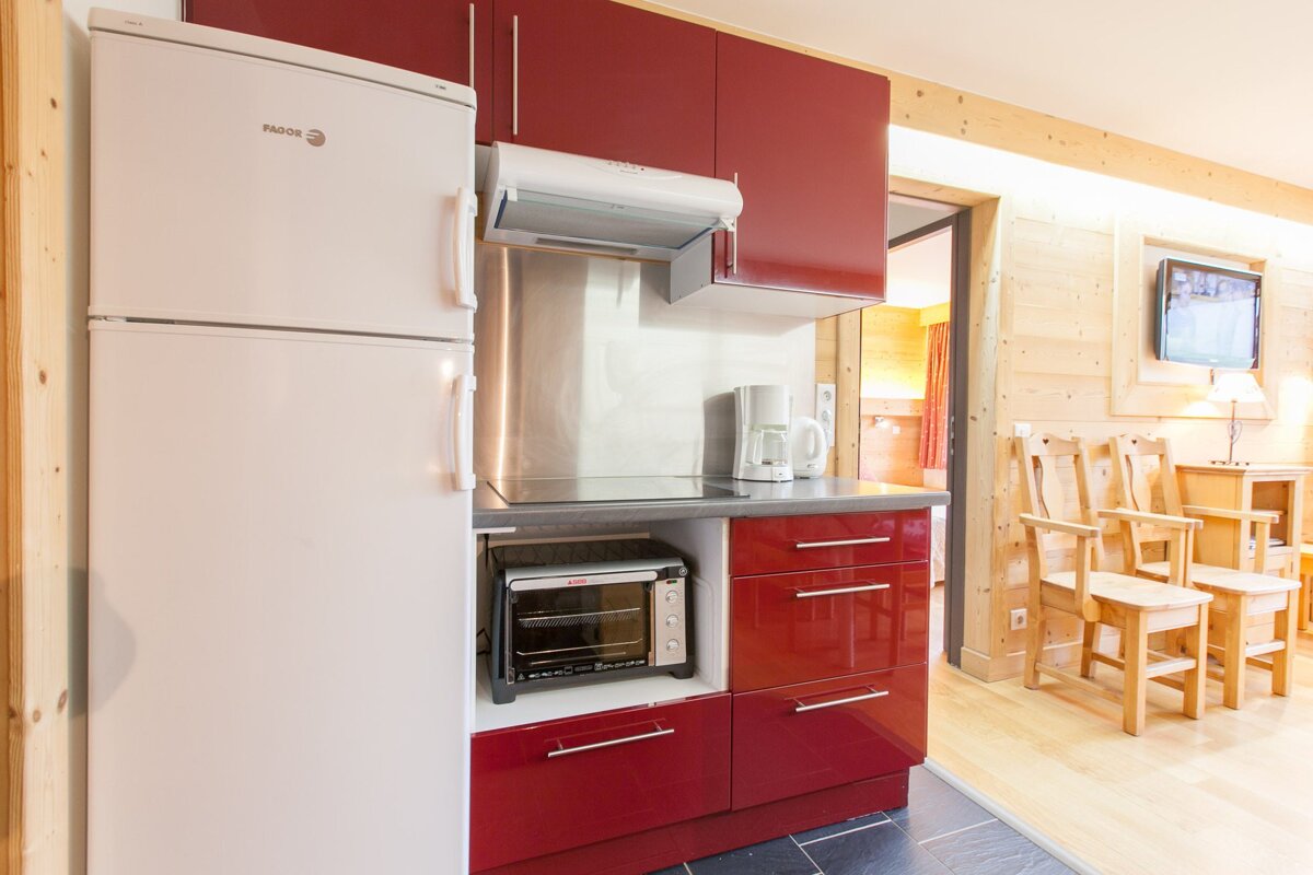 A compact kitchen with red cabinets and a white fridge, integrated into a wood-paneled room featuring wooden chairs and a TV in the background.