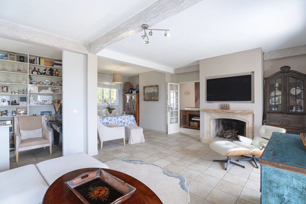 An elegant living room with a white sofa, fireplace, TV, and bookshelves, flowing into a dining area with a set table, all with tiled floors.
