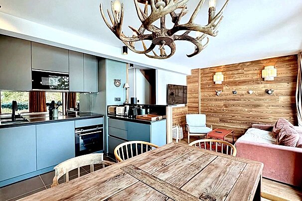 An open concept, modern chalet-style space with a large antler chandelier, teal kitchen, warm wood paneled walls, and a rustic dining table.