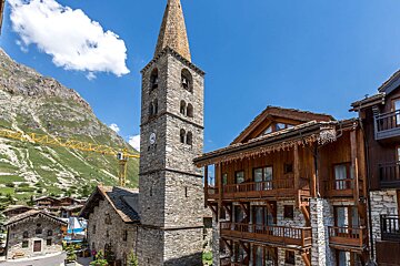 A stone church tower dominates an alpine village with wooden chalets, set against green mountains and a blue sky. A construction crane is visible.