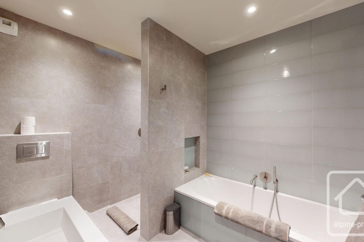 A modern bathroom with light grey tiled walls, featuring a walk-in shower, a bathtub with a towel, and a toilet. Recessed lighting brightens the space.