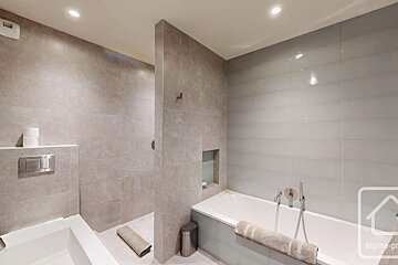 A modern bathroom with light grey tiled walls, featuring a walk-in shower, a bathtub with a towel, and a toilet. Recessed lighting brightens the space.
