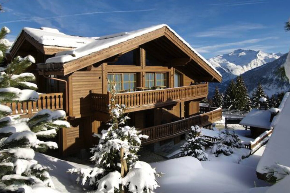 A beautiful wooden chalet in a snow-covered mountain landscape with pine trees and a clear blue sky.