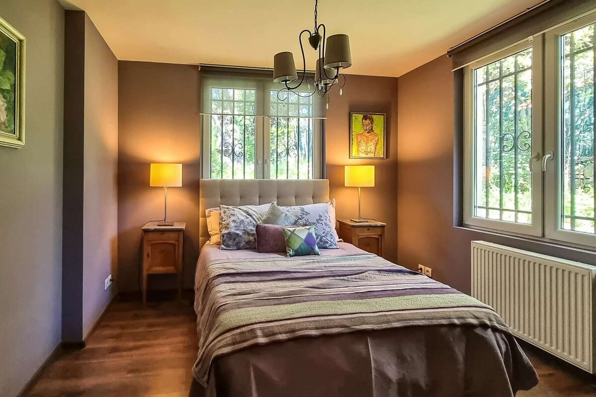 A cozy bedroom features a bed with nightstands and lamps, windows with decorative grilles, warm brown walls, and a central chandelier.
