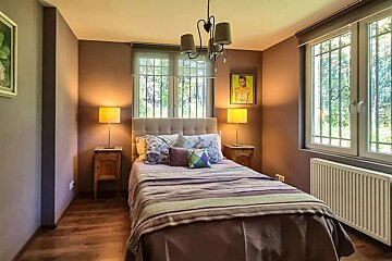 A cozy bedroom features a bed with nightstands and lamps, windows with decorative grilles, warm brown walls, and a central chandelier.