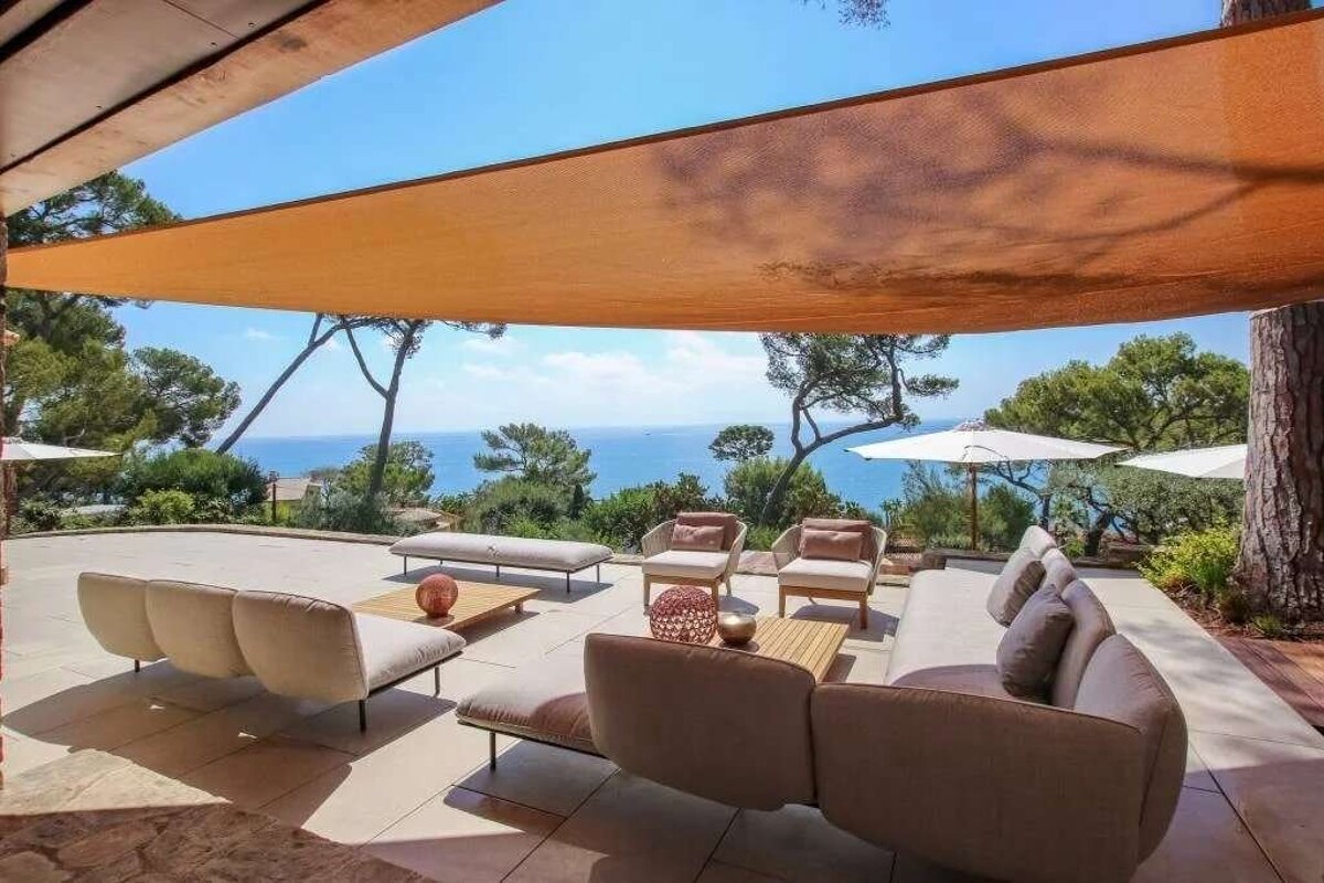 Stylish outdoor patio with modern seating under a shade sail, offering stunning ocean views framed by trees on a sunny day.