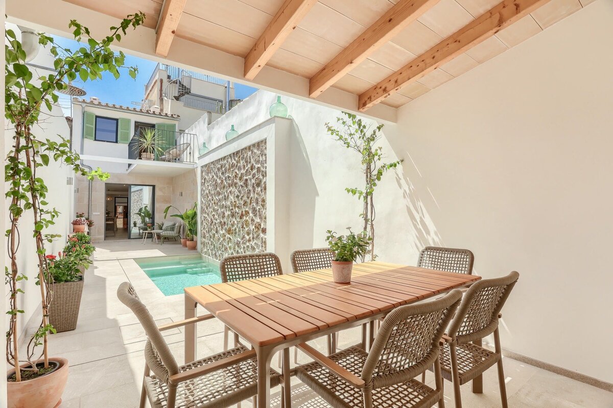 An inviting outdoor patio with a dining table, chairs, a small pool, and a white building with green shutters under a sunny sky.