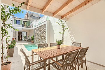 A sunny Mediterranean courtyard featuring an outdoor dining set under a wooden pergola, a small pool, and white walls with stone accents and climbing plants.