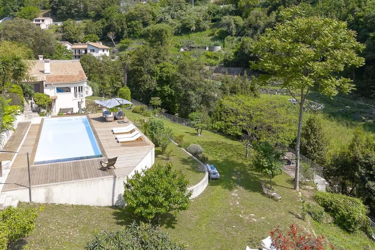 Aerial view of a white house with a pool and wooden deck, nestled on a lush, green hillside with numerous trees and distant buildings.