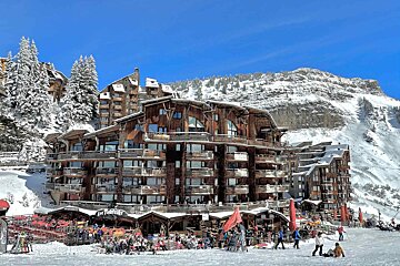 Sunny ski resort with large wooden chalets, snow-covered mountains, and people enjoying outdoor dining and activities on the slopes.