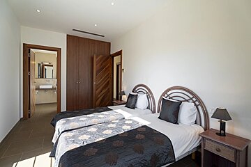 Bright twin bedroom with dark patterned bedding, rattan headboards, a wooden wardrobe, and an open door revealing an ensuite bathroom.
