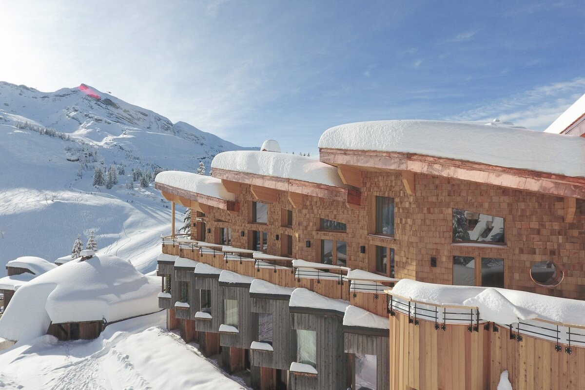 Modern ski resort with curved, snow-covered roofs and wood shingle facade, nestled in snowy mountains under a clear sky.