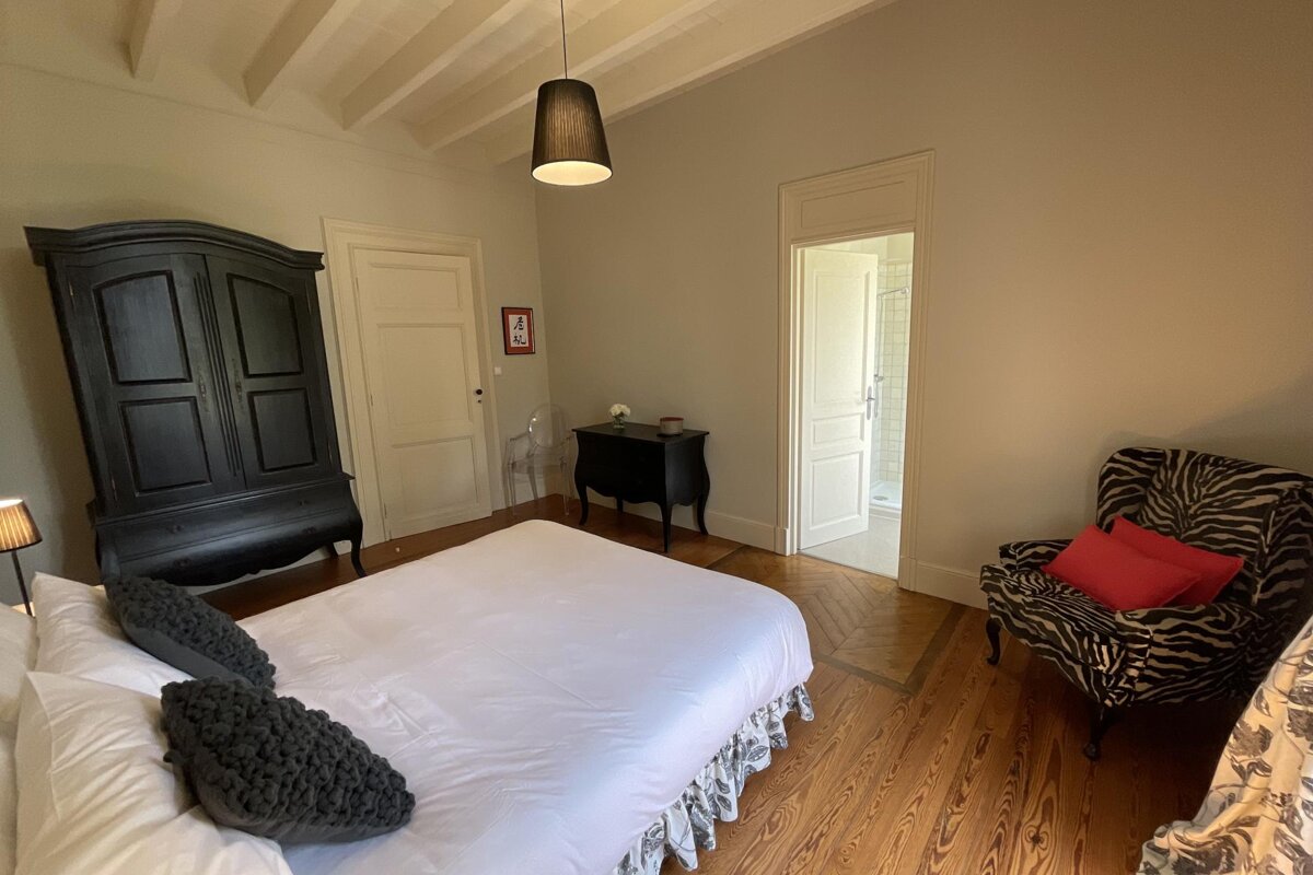 A bright bedroom with a white bed, black armoire, and a zebra print armchair with red pillows. Features wooden floors and light walls, with a bathroom visible.