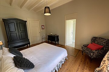 A bright bedroom with a white bed, black armoire, and a zebra print armchair with red pillows. Features wooden floors and light walls, with a bathroom visible.