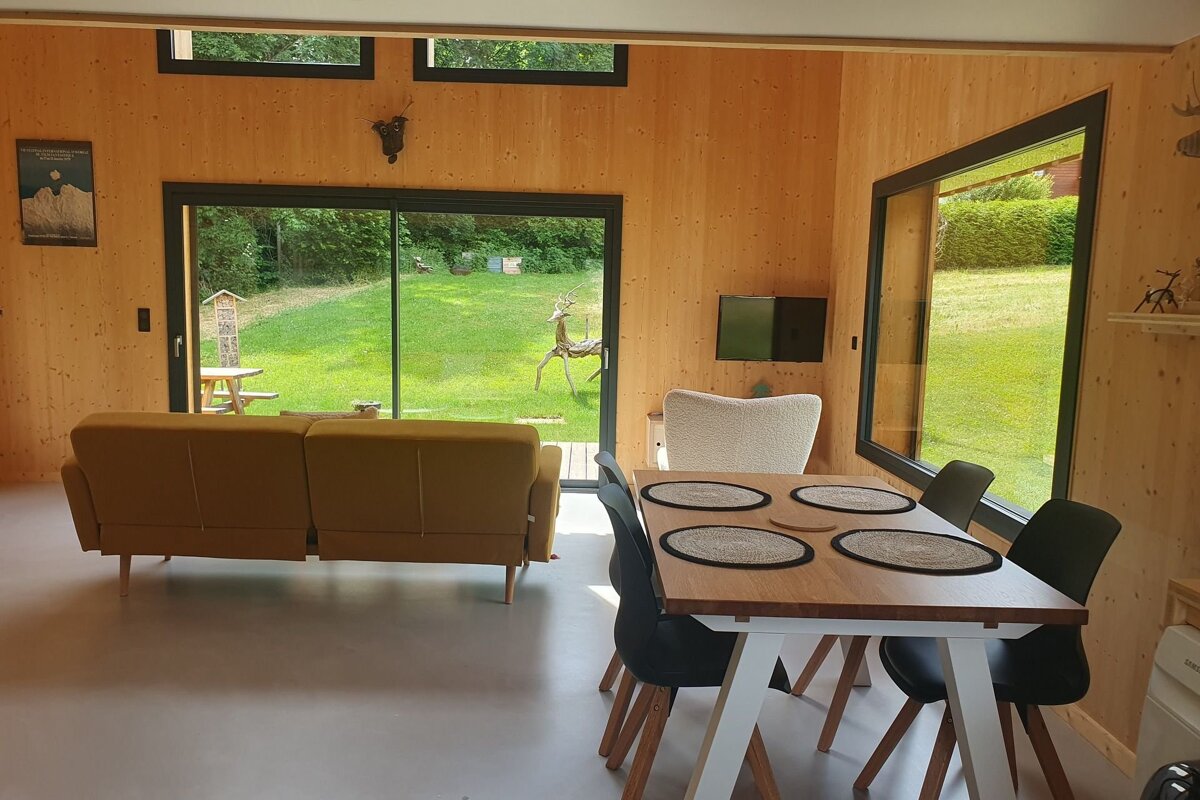 Cozy, wood-paneled room with a sofa and dining table. Large windows provide a view of a sunny green lawn with a deer statue and a picnic table.